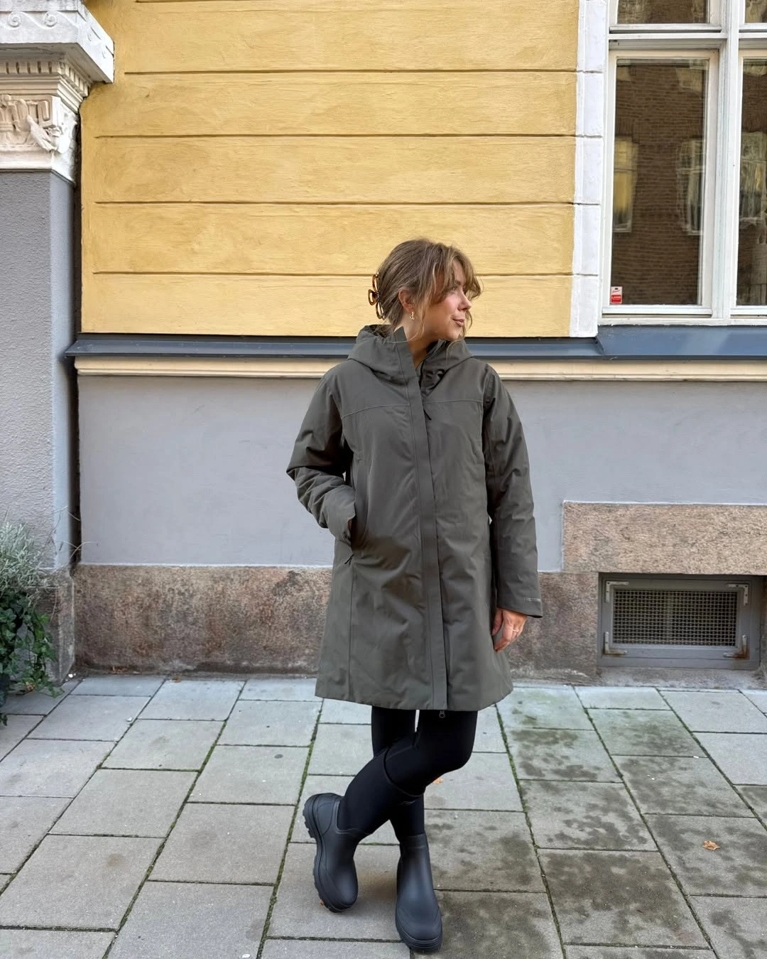 Olive-gray hooded coat with black fitted pants and dark ankle boots