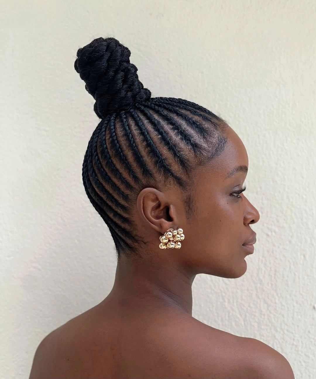 Cornrows pulled upward into tall braided bun with gold earrings