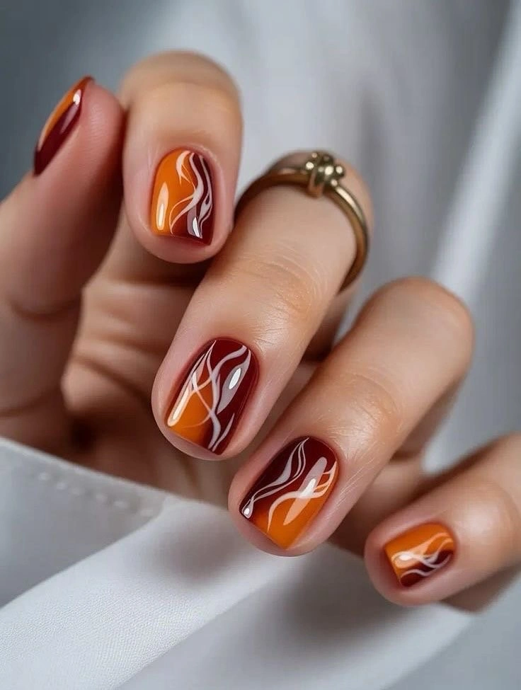 Short maroon and orange nails with white swirl accents