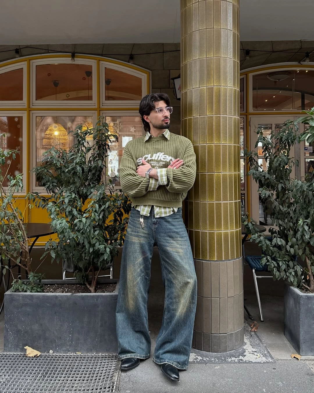 Man in olive graphic sweater, plaid shirt and very wide jeans